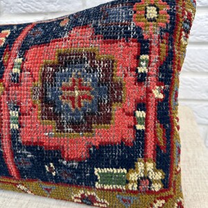 May include: A rectangular pillow with a detailed, vintage rug design. The pillow features a central pattern in shades of red and blue, with accents of gold and green. The textured surface suggests a woven or embroidered construction.