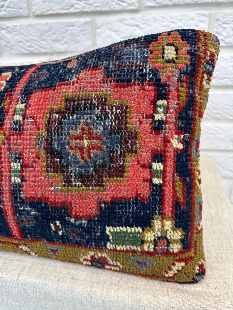 May include: A rectangular pillow with a detailed, vintage rug design. The pillow features a central pattern in shades of red and blue, with accents of gold and green. The textured surface suggests a woven or embroidered construction.