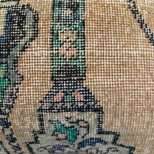 May include: Close-up of a rectangular cushion with a vintage, patterned design. The cushion features a beige background with intricate geometric patterns in shades of blue, green, black, and pink. The texture appears woven.