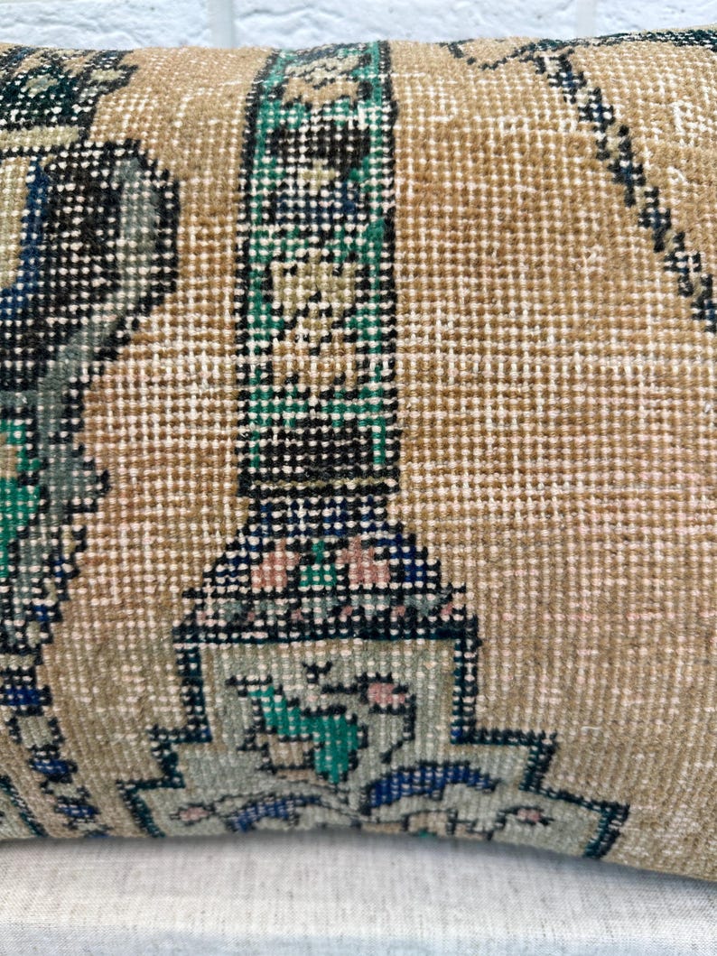 May include: Close-up of a rectangular cushion with a vintage, patterned design. The cushion features a beige background with intricate geometric patterns in shades of blue, green, black, and pink. The texture appears woven.