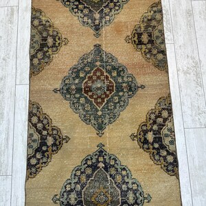 May include: A long, rectangular runner rug featuring a tan background with a repeating pattern of large, ornate diamond-shaped medallions in shades of dark blue, muted green, and deep red, with intricate floral-inspired geometric details.