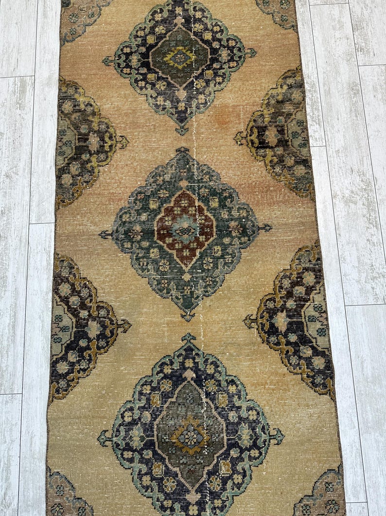 May include: A long, rectangular runner rug featuring a tan background with a repeating pattern of large, ornate diamond-shaped medallions in shades of dark blue, muted green, and deep red, with intricate floral-inspired geometric details.