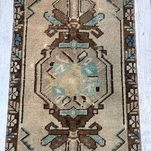 May include: A rectangular, vintage rug with a central medallion design. The rug has a beige background with brown, teal, and cream geometric patterns. The border features a repeating floral motif.
