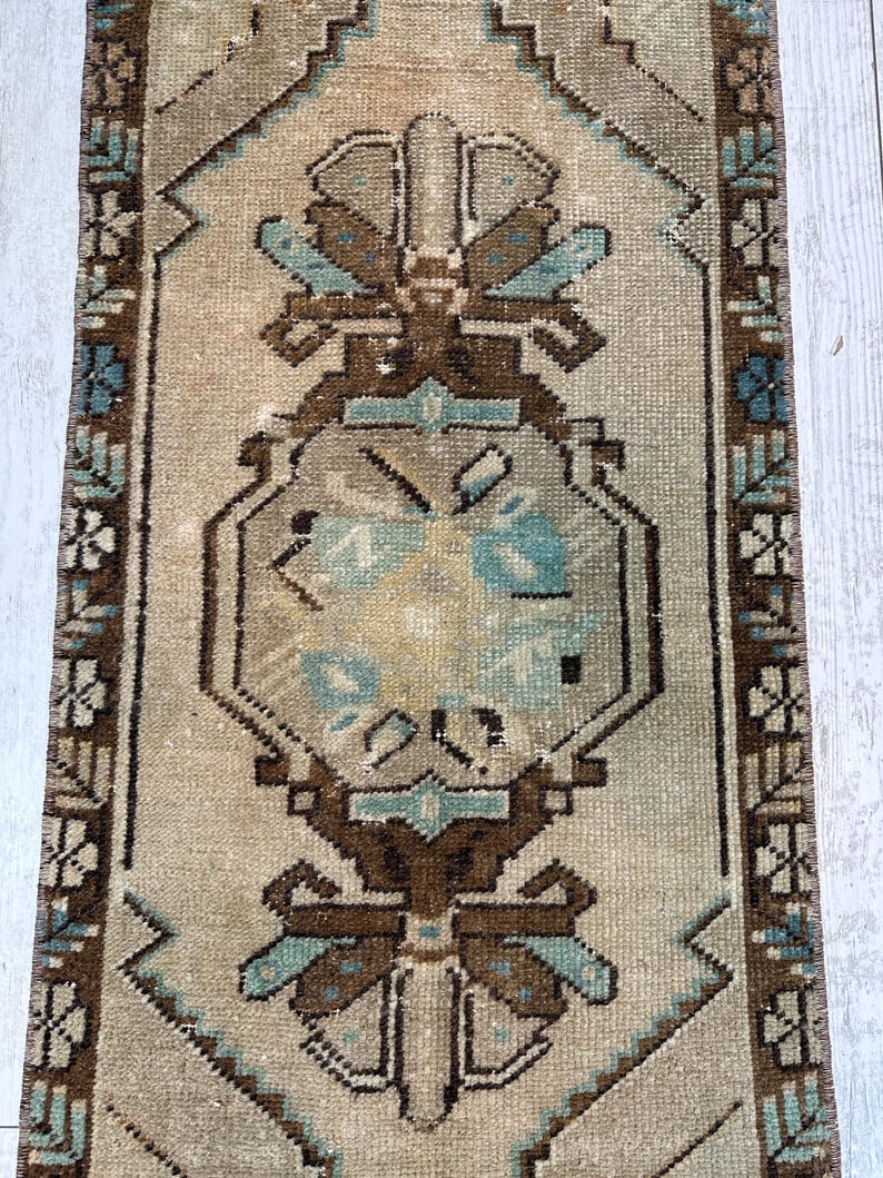May include: A rectangular, vintage rug with a central medallion design. The rug has a beige background with brown, teal, and cream geometric patterns. The border features a repeating floral motif.