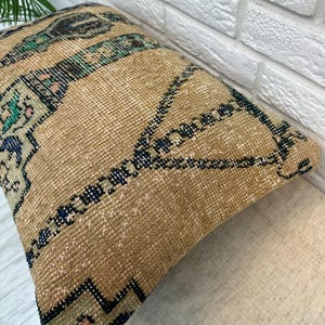 May include: A rectangular, beige cushion with a geometric pattern in shades of blue, green, and black. The cushion has a textured, woven appearance, suggesting a vintage or handcrafted style. The design features repeating shapes and lines.