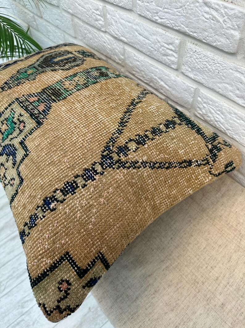May include: A rectangular, beige cushion with a geometric pattern in shades of blue, green, and black. The cushion has a textured, woven appearance, suggesting a vintage or handcrafted style. The design features repeating shapes and lines.