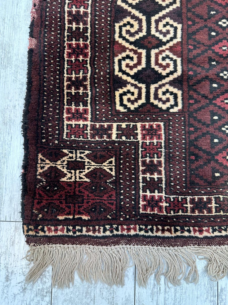 May include: Close-up of a section of a burgundy and cream-colored rug with geometric patterns. The rug has a fringe of beige tassels along the bottom edge. The design features repeating shapes and borders.
