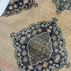May include: A woven rug featuring a light tan base with repeating ornate medallion patterns in shades of dark blue, teal, mustard yellow, and brown. The design includes intricate geometric and floral-inspired motifs throughout.