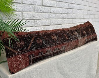 May include: A long, rectangular lumbar pillow featuring a woven textile cover with a dark red and black geometric pattern. The fabric displays a distressed, aged texture with intricate border details and repeating motifs across the entire surface.