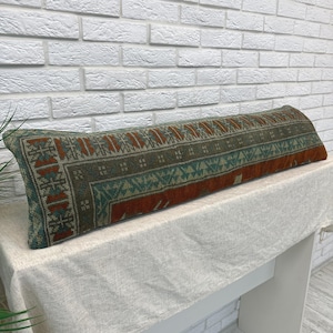 May include: A long, rectangular lumbar pillow featuring a woven textile cover with a geometric pattern in shades of rust orange, muted teal, and dark olive green. The fabric displays a dense, intricate design with repeating motifs and borders.