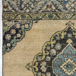 May include: A woven rug featuring a tan background with a central medallion and corner motifs in shades of dark brown, teal, light blue, and olive green. The border displays a geometric pattern in blue, tan, and green tones.