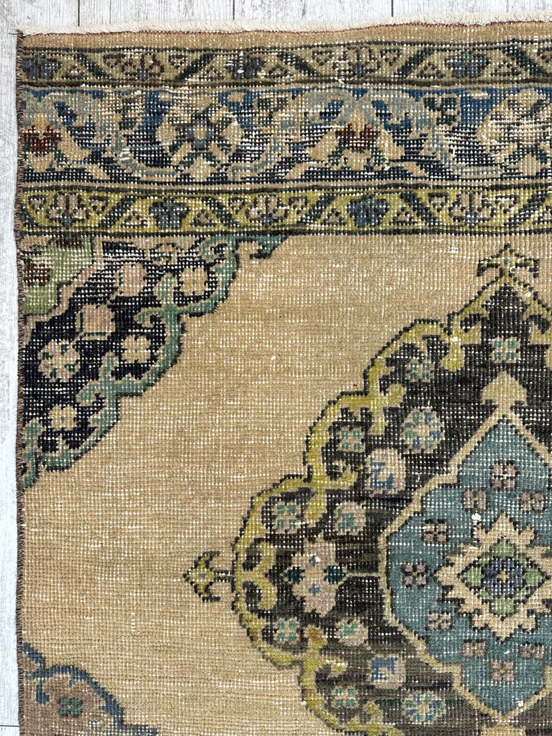 May include: A woven rug featuring a tan background with a central medallion and corner motifs in shades of dark brown, teal, light blue, and olive green. The border displays a geometric pattern in blue, tan, and green tones.