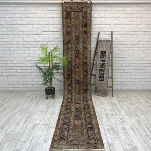 May include: A long, narrow runner rug featuring a dark brown and muted olive green geometric pattern with faded orange and teal floral accents. The rug hangs vertically, displaying a symmetrical design with a central border and intricate motifs.