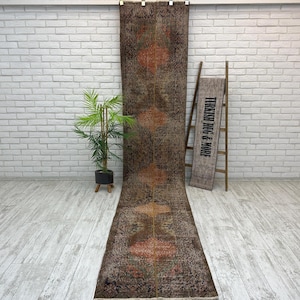 May include: A long, narrow runner rug featuring a dark brown base with repeating geometric diamond-shaped motifs in shades of orange and muted green. The rug displays a weathered, distressed texture throughout its surface, with a thin vertical line.