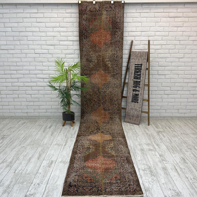 May include: A long, narrow runner rug featuring a dark brown base with repeating geometric diamond-shaped motifs in shades of orange and muted green. The rug displays a weathered, distressed texture throughout its surface, with a thin vertical line.