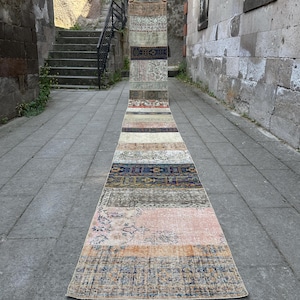 Op de afbeelding: Een lange, smalle loper met een patchworkontwerp bestaande uit diverse rechthoekige secties. Het kleed toont een gevarieerd kleurenpalet met tinten blauw, oranje, roze, beige en bruin, voorzien van ingewikkelde patronen en texturen.