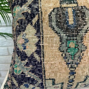 May include: A square cushion with a vintage rug design. The cushion has a beige background with geometric patterns in navy blue, teal, and pink. The cushion is made of a woven fabric.