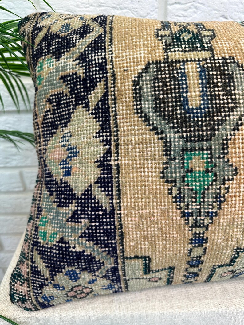 May include: A square cushion with a vintage rug design. The cushion has a beige background with geometric patterns in navy blue, teal, and pink. The cushion is made of a woven fabric.
