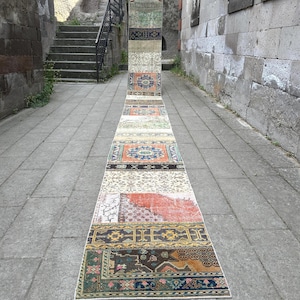 Può includere: Un tappeto passatoia lungo e stretto caratterizzato da un design patchwork composto da varie sezioni rettangolari. Il tappeto presenta una tavolozza di colori variegata che include tonalità arancione, blu, verde, giallo, marrone e crema, con intricati motivi floreali e geometrici.