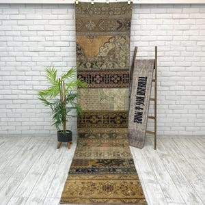 May include: A long, rectangular, patchwork rug with a variety of patterns and colors, including beige, brown, and navy. The rug is hanging vertically against a white brick wall. A wooden ladder and a sign that reads "TURKISH RUG & MORE" are leaning against the wall.
