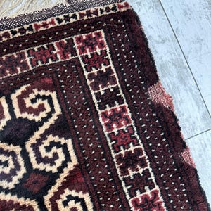 May include: Close-up of a section of a hand-woven rug. The rug features a deep burgundy and cream color palette with geometric patterns. The edges have a fringe detail, adding texture and visual interest to the rug.