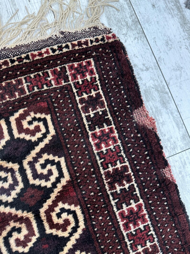 May include: Close-up of a section of a hand-woven rug. The rug features a deep burgundy and cream color palette with geometric patterns. The edges have a fringe detail, adding texture and visual interest to the rug.