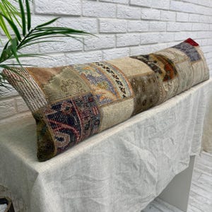 May include: A long, rectangular cushion with a patchwork design. The cushion features a variety of colours and patterns, including beige, brown, blue, and red. The cushion is resting on a white surface.