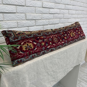 May include: A long, rectangular pillow with a traditional rug design. The pillow features a deep red background with intricate floral patterns in shades of blue, yellow, and beige. The pillow is placed on a white surface against a white brick wall.