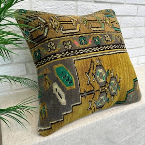May include: A square throw cushion featuring a textured, woven surface with a multi-coloured geometric pattern in shades of mustard yellow, charcoal grey, and teal green. The design includes intricate borders and central floral-like motifs.