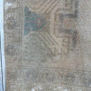 K&ouml;nnte beinhalten: Ein gewebter Teppich in beigen und hellbraunen Farbt&ouml;nen mit verblassten geometrischen Mustern in sanftem Grau und T&uuml;rkis. Das Textil weist eine abgenutzte Struktur mit sichtbarem Flor und einer kurzen wei&szlig;en Franse an der unteren Kante auf.