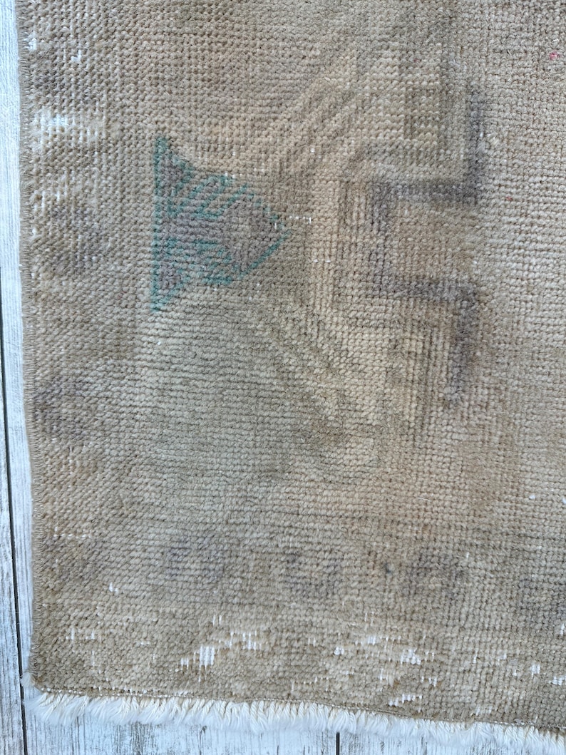K&ouml;nnte beinhalten: Ein gewebter Teppich in beigen und hellbraunen Farbt&ouml;nen mit verblassten geometrischen Mustern in sanftem Grau und T&uuml;rkis. Das Textil weist eine abgenutzte Struktur mit sichtbarem Flor und einer kurzen wei&szlig;en Franse an der unteren Kante auf.
