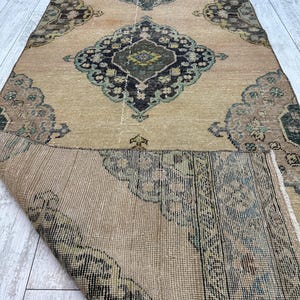 May include: A long rectangular runner rug featuring a tan base color with repeating dark blue, green, and brown geometric medallion patterns. The rug is partially folded over, revealing the woven texture and underside of the fabric.