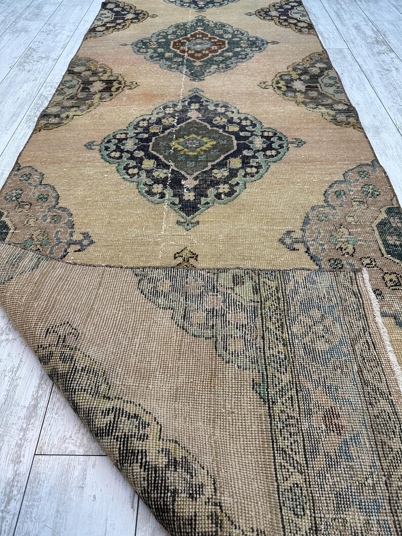 May include: A long rectangular runner rug featuring a tan base color with repeating dark blue, green, and brown geometric medallion patterns. The rug is partially folded over, revealing the woven texture and underside of the fabric.