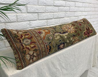 Peut inclure: Un long coussin traversin rectangulaire présentant un motif tissé détaillé dans des tons vert olive, beige, rouge foncé et crème. Le textile affiche des motifs floraux et géométriques complexes sur toute sa surface, avec un bord passepoilé.