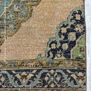 May include: A woven rug featuring a tan background with a large central medallion in shades of teal, olive green, and dark blue. The border displays a repeating floral pattern in dark blue, teal, and olive green with a fringed edge.