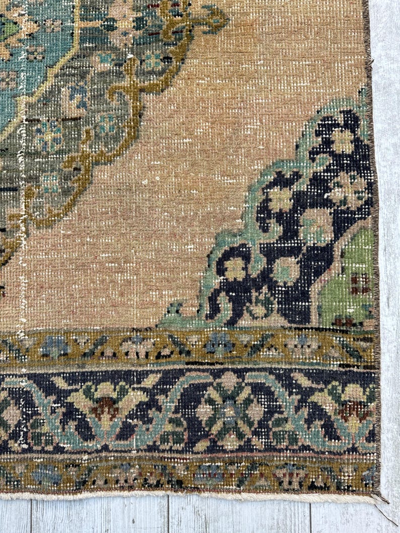 May include: A woven rug featuring a tan background with a large central medallion in shades of teal, olive green, and dark blue. The border displays a repeating floral pattern in dark blue, teal, and olive green with a fringed edge.
