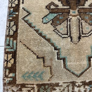May include: Close-up of a beige and brown vintage rug with a geometric pattern. The rug features a central design with brown and teal accents, surrounded by a detailed border. The rug has a fringed edge and a textured appearance.