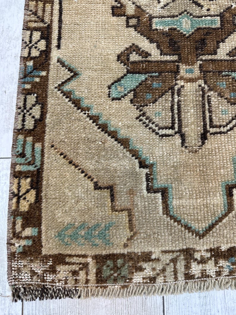 May include: Close-up of a beige and brown vintage rug with a geometric pattern. The rug features a central design with brown and teal accents, surrounded by a detailed border. The rug has a fringed edge and a textured appearance.