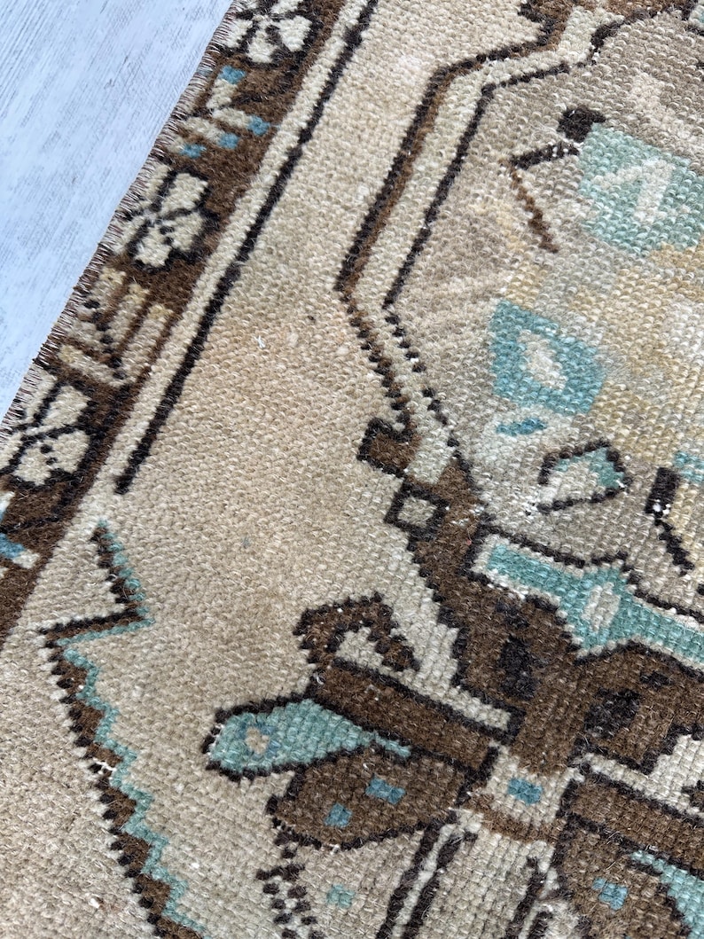 May include: Close-up of a beige and brown patterned rug with geometric and floral designs. The rug features accents of light blue and a textured surface, showcasing a traditional style.