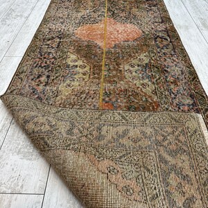 May include: A long, rectangular runner rug featuring a complex, distressed pattern in shades of brown, muted orange, and dark blue. The rug has a central vertical yellow line and a folded corner revealing the woven, textured underside of the fabric.