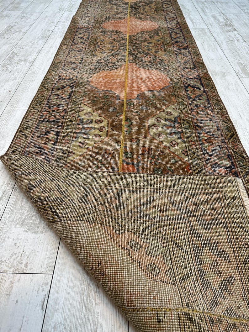 May include: A long, rectangular runner rug featuring a complex, distressed pattern in shades of brown, muted orange, and dark blue. The rug has a central vertical yellow line and a folded corner revealing the woven, textured underside of the fabric.