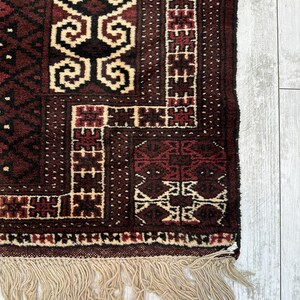 May include: Close-up of a section of a hand-woven rug. The rug features a complex geometric pattern in shades of burgundy, black, and cream. The edges are finished with a fringe of natural-colored fibers, adding texture and detail to the design.