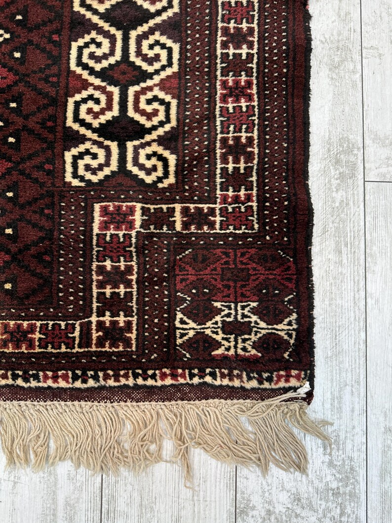 May include: Close-up of a section of a hand-woven rug. The rug features a complex geometric pattern in shades of burgundy, black, and cream. The edges are finished with a fringe of natural-colored fibers, adding texture and detail to the design.