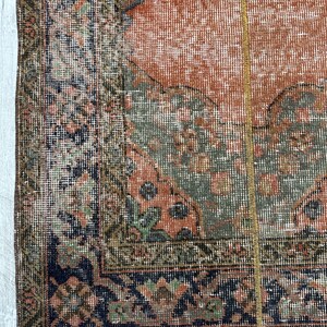 May include: A woven rug featuring a central terracotta orange field with sage green and dark blue geometric patterns. The border displays intricate floral and diamond motifs in navy, olive, and muted pink tones, finished with a short white fringe.