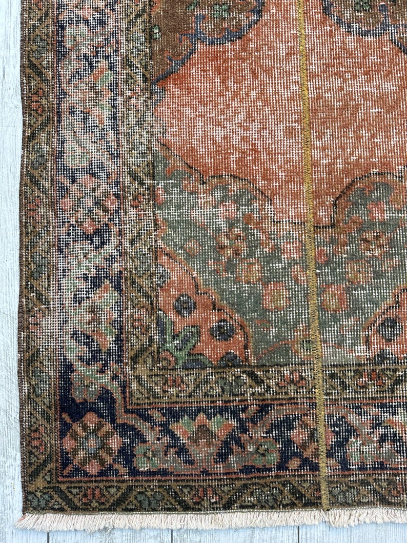 May include: A woven rug featuring a central terracotta orange field with sage green and dark blue geometric patterns. The border displays intricate floral and diamond motifs in navy, olive, and muted pink tones, finished with a short white fringe.