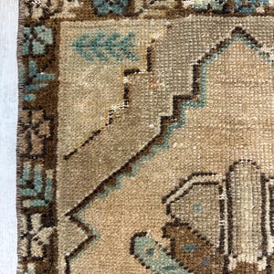 May include: Close-up of a section of a vintage rug in beige and brown tones. The rug displays geometric and floral patterns in brown and teal. The edges are fringed.