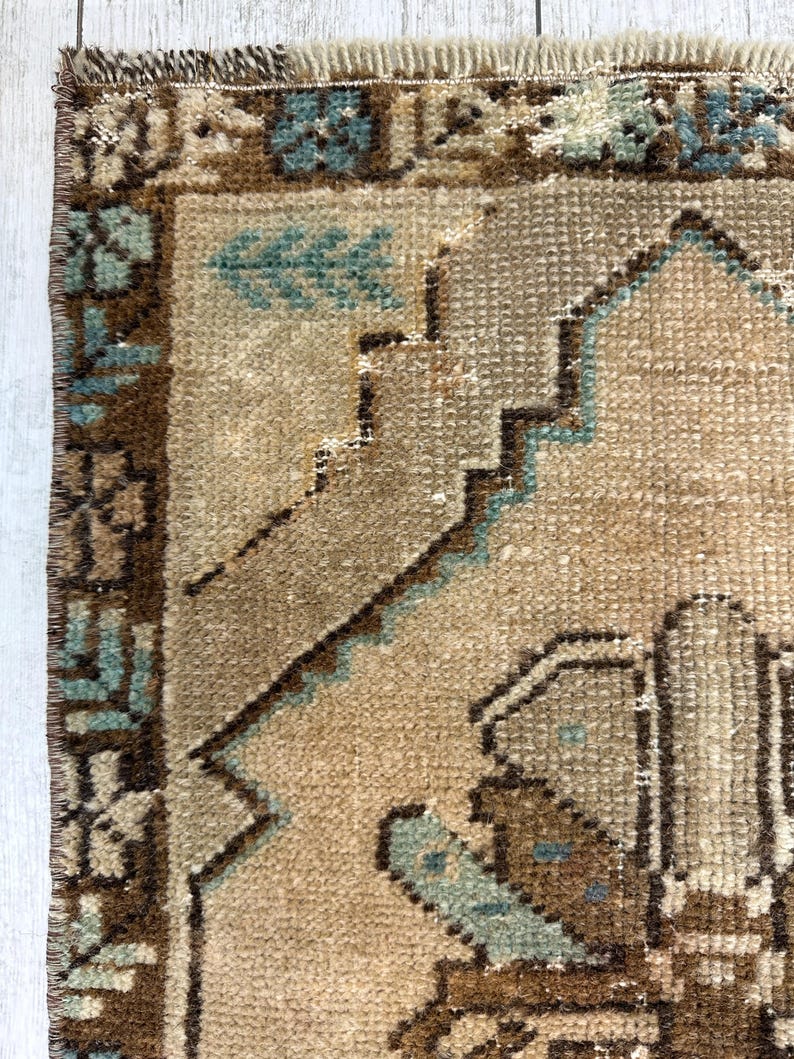 May include: Close-up of a section of a vintage rug in beige and brown tones. The rug displays geometric and floral patterns in brown and teal. The edges are fringed.