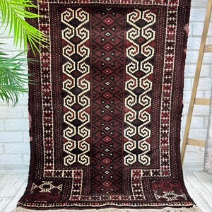 May include: A rectangular, dark brown rug with a central design of repeating cream-colored scroll-like shapes and diamond patterns. The rug has a border of red and cream geometric patterns and cream fringe along the bottom edge. The rug is hanging on a wall.