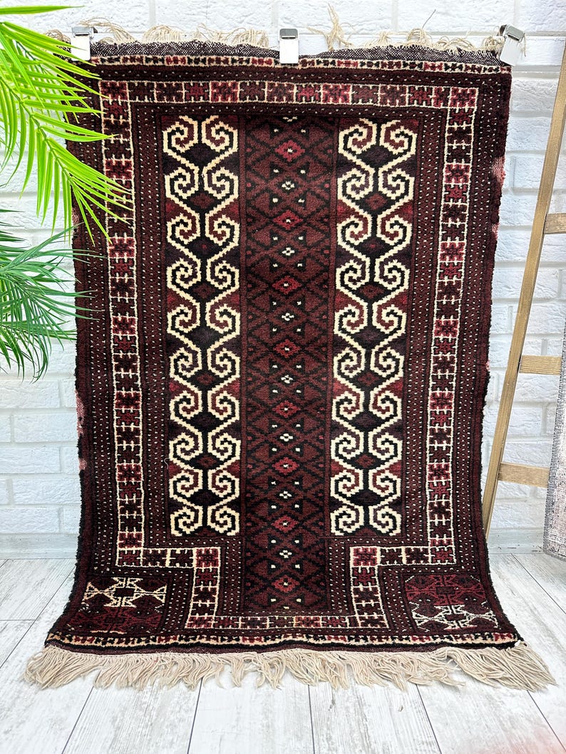 May include: A rectangular, dark brown rug with a central design of repeating cream-colored scroll-like shapes and diamond patterns. The rug has a border of red and cream geometric patterns and cream fringe along the bottom edge. The rug is hanging on a wall.