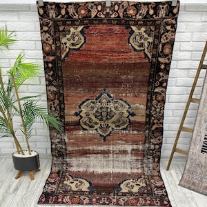 Puede incluir: Una alfombra rectangular, desgastada, con un diseño de medallón central. La alfombra presenta una paleta de colores rojo, marrón y beige, con un borde oscuro. La alfombra está colgada contra una pared de ladrillos blancos.